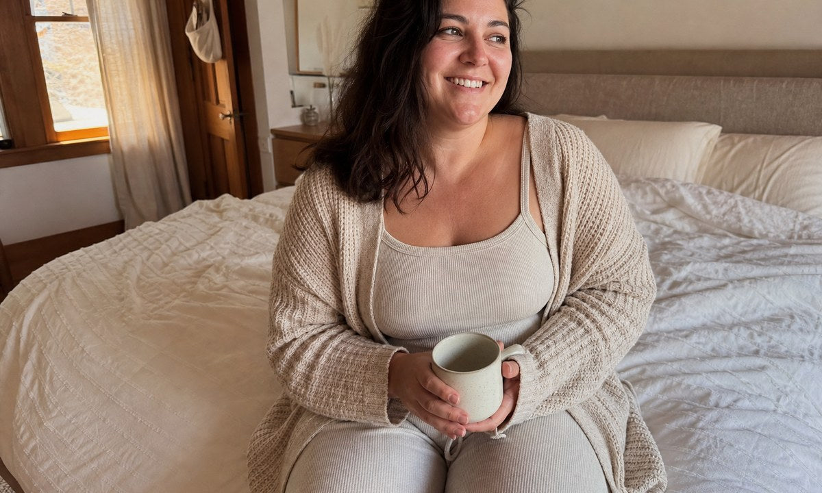 Woman in oat ribbed loungewear and knit cardigan sitting on a linen bed with a mug in warm morning light